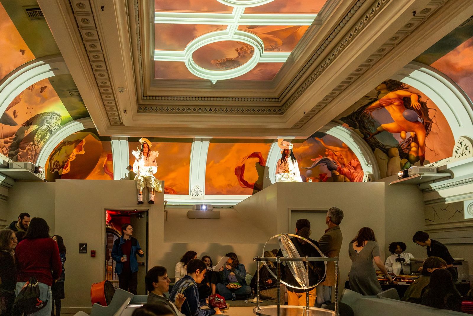 Two priestesses sit above a crowd underneath a brightly colored mural made interactive with glowing projection.